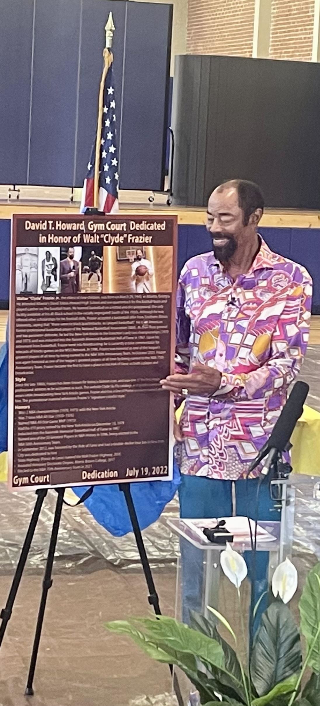Walt Frazier standing next to dedication poster of the gym being dedicated to him.